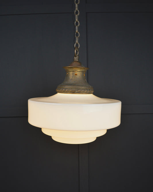 A vintage-style pendant light with a tiered white glass shade and an antique brass top cap, hanging from a chain against a dark paneled wall.