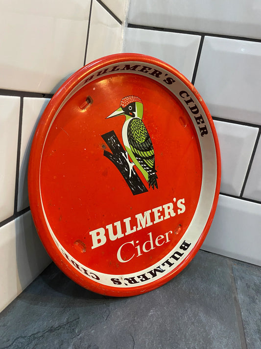Vintage Bulmer's Cider metal tray with woodpecker graphic, red and white, against tiled wall.