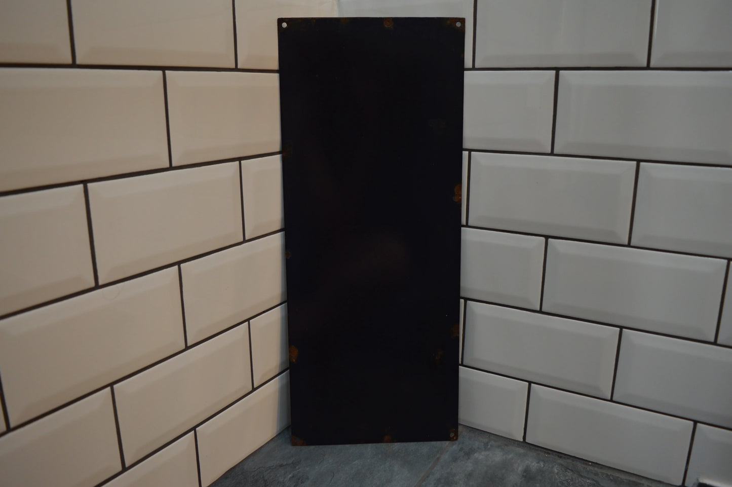 Vintage black metal sign panel with rust spots leaning against white subway tiles