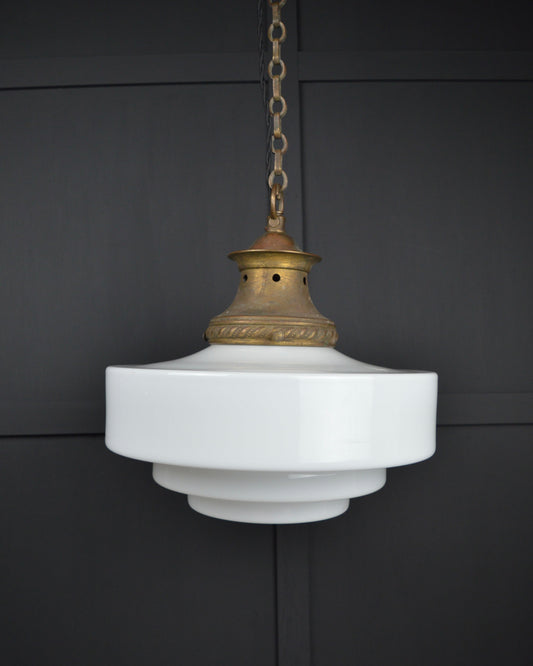 A vintage-style pendant light with a tiered white glass shade and an ornate brass top cap, suspended by a chain against a dark paneled wall.
