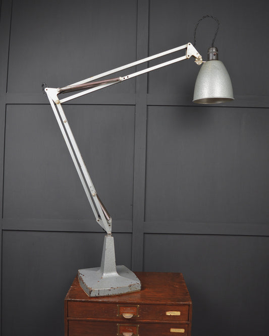 A vintage-style adjustable desk lamp with a white articulated arm and a silver dome shade, resting on a brown wooden drawer cabinet against a dark gray paneled wall.