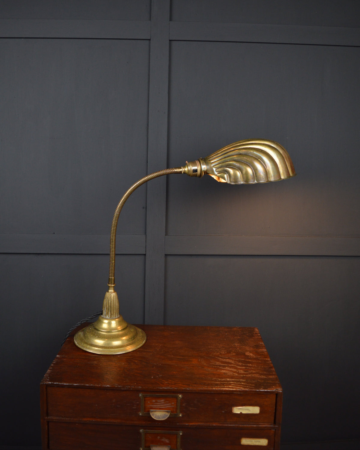 A brass desk lamp with a curved gooseneck and a fluted, fan-like shade, displayed on a vintage wooden chest against a dark paneled wall.