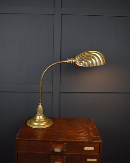 A brass desk lamp with a curved gooseneck and a fluted, fan-like shade, displayed on a vintage wooden chest against a dark paneled wall.